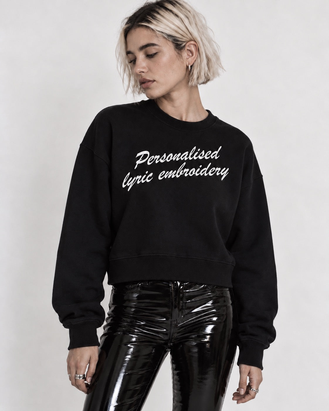 A woman wearing a black crewneck sweatshirt with white cursive text reading 'Personalised lyric embroidery' and shiny black pants, photographed against a plain light gray background.