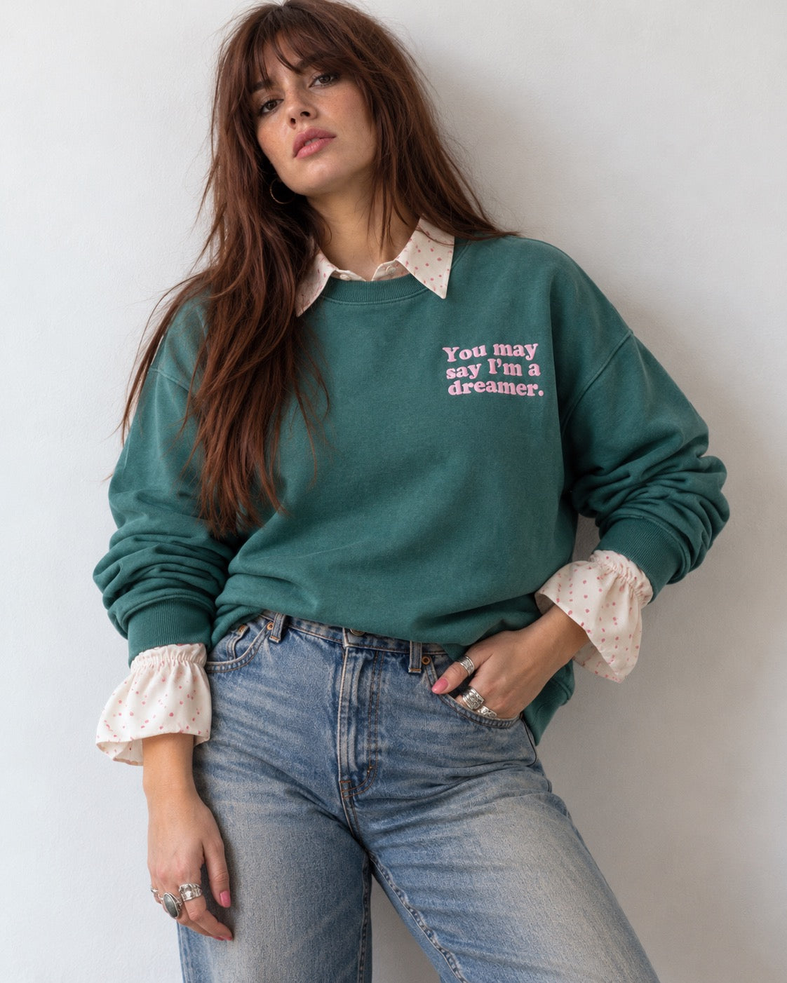 A woman wearing a sage green crewneck sweatshirt with a pink text graphic that reads 'You may say I'm a dreamer.' on the left chest, styled over a cream ruffled blouse and blue jeans.