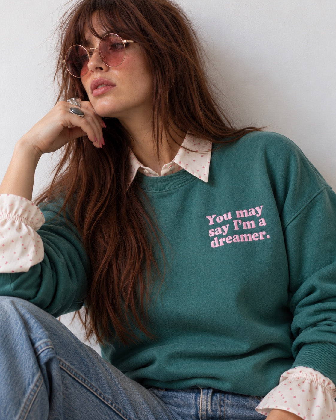 A woman wearing a dark teal green sweatshirt with pink embroidered text that reads 'You may say I'm a dreamer.' She is styled with a white polka-dot collared shirt underneath, blue jeans, and round sunglasses, posing against a plain light gray wall.
