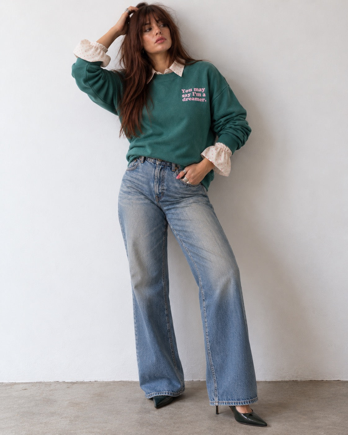 A woman models a dark green crewneck sweatshirt with white graphic text on the chest, layered over a white ruffled blouse, paired with wide-leg blue jeans and black pointed-toe heels, standing against a plain white wall.