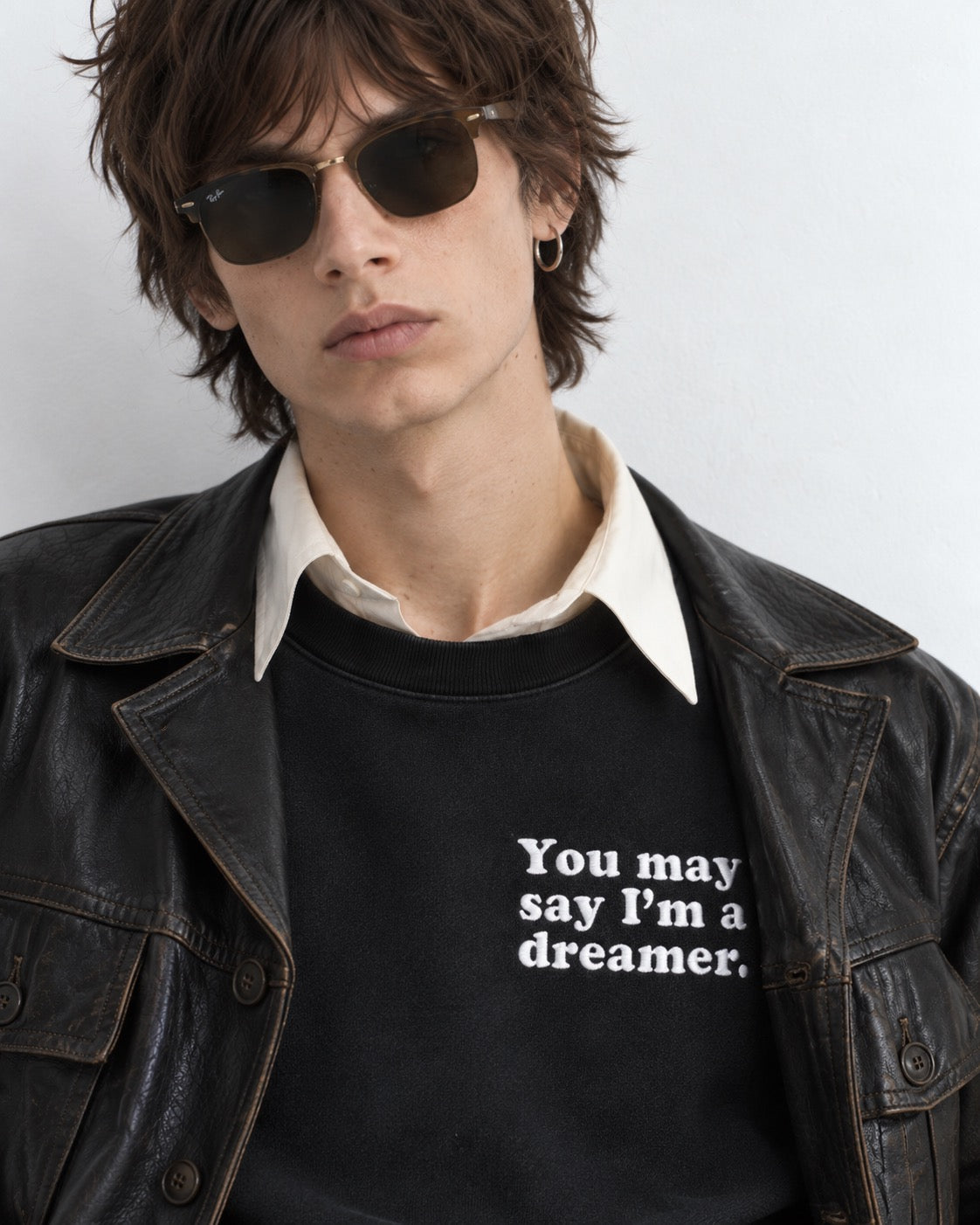 A model wearing a black crewneck sweatshirt with the white printed text 'You may say I'm a dreamer.' layered under an open black leather jacket and a cream collared shirt, accessorized with dark sunglasses.